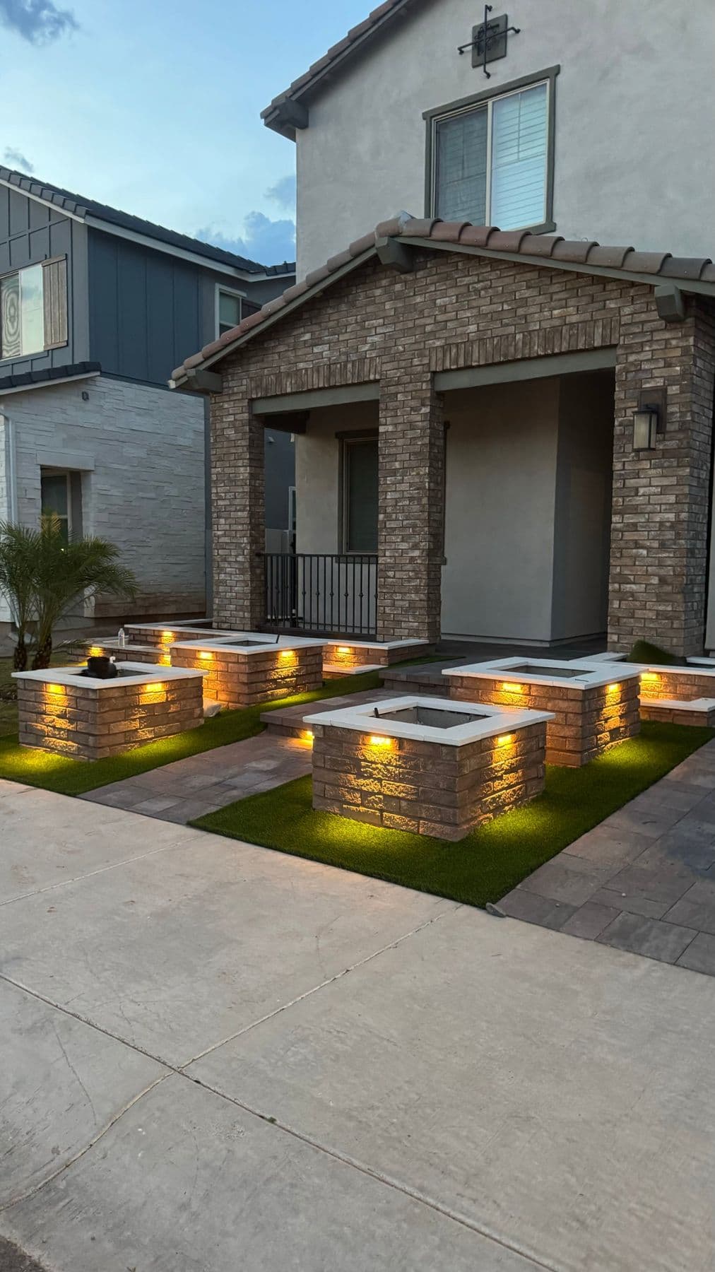 Front yard at dusk with lit masonry planters, paver walkways and artificial turf
