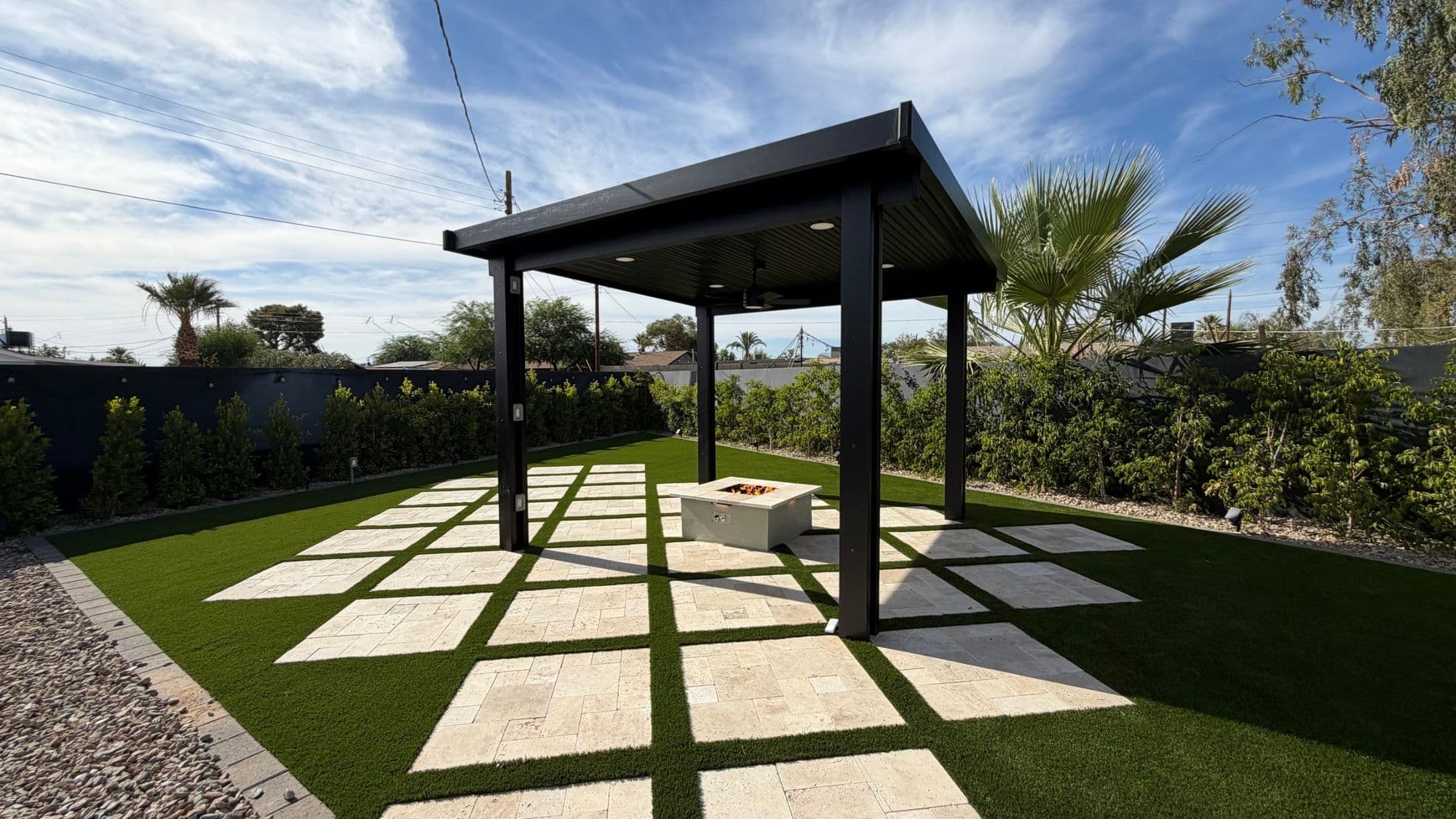 Black aluminum pergola over ivory travertine pads and a propane fire pit, set in artificial turf