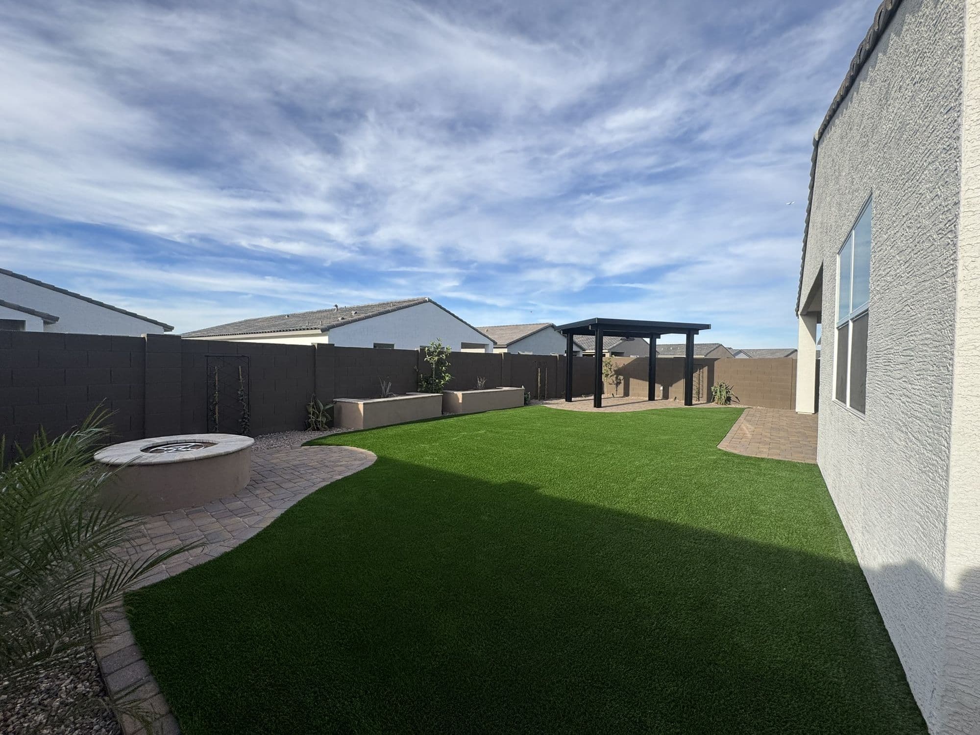 Backyard with curved artificial turf, paver patios, a round gas fire pit, raised planters and a black aluminum pergola
